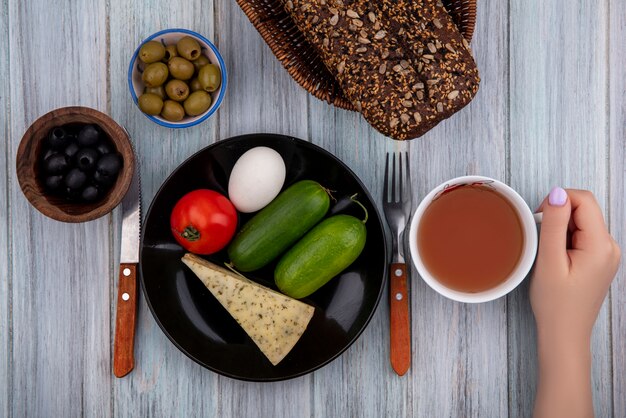 Vista superior mujer sostenga una taza de té con queso pepinos tomate y huevo en un plato con aceitunas negras y verdes sobre un fondo gris