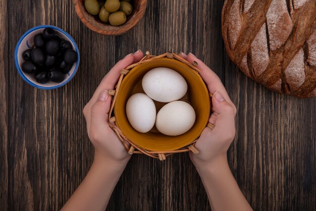 Vista superior mujer sosteniendo huevos de gallina en canasta con aceitunas y pan negro sobre fondo de madera