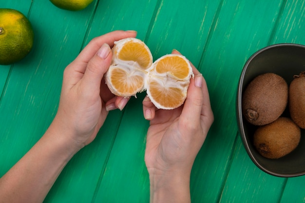 Vista superior mujer sosteniendo mandarina pelada en sus manos con kiwi en un recipiente sobre una pared verde