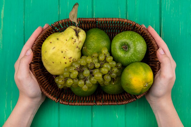 Vista superior mujer sosteniendo manzanas verdes con pera, mandarinas y uvas en una canasta en una pared verde