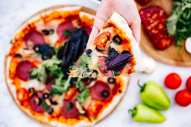 Vista superior de la mujer sosteniendo una rebanada de pizza de pepperoni con champiñones y hierbas de tomate verde oliva