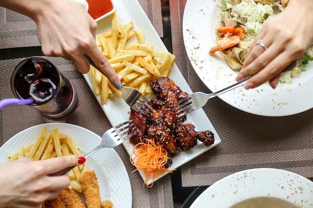 Vista superior mujeres comiendo alitas de pollo a la barbacoa con papas fritas y ensalada con jugo sobre la mesa