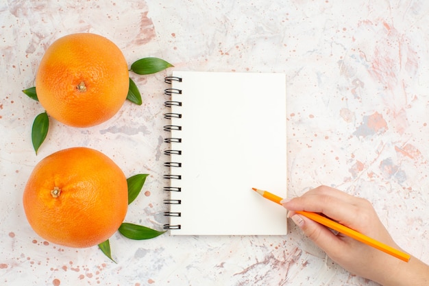 Vista superior de naranjas frescas cuaderno naranja lápiz en mano femenina en superficie aislada brillante con espacio de copia