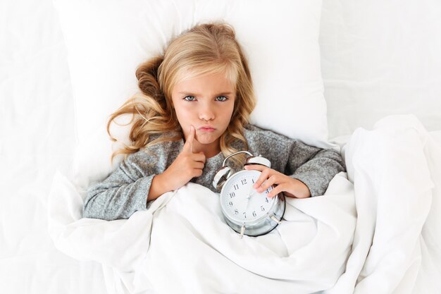 Vista superior de la niña de pensamiento infeliz tocando su rostro con el dedo mientras está acostado en la cama con reloj despertador,