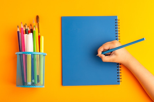 Vista superior del niño pequeño dibujando y escribiendo algo en un cuaderno sobre la superficie naranja