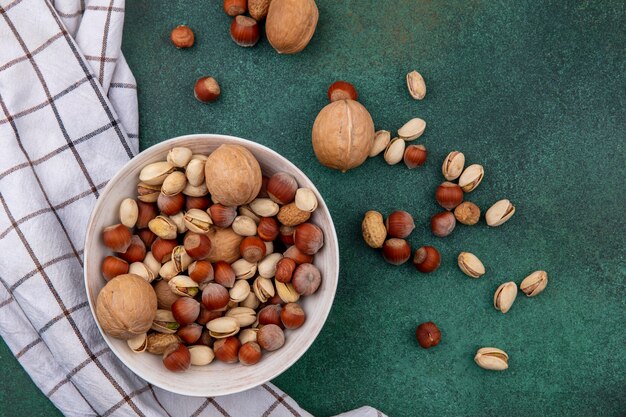 Vista superior de nueces con avellanas, pistachos y cacahuetes en un recipiente con una toalla a cuadros sobre una mesa verde