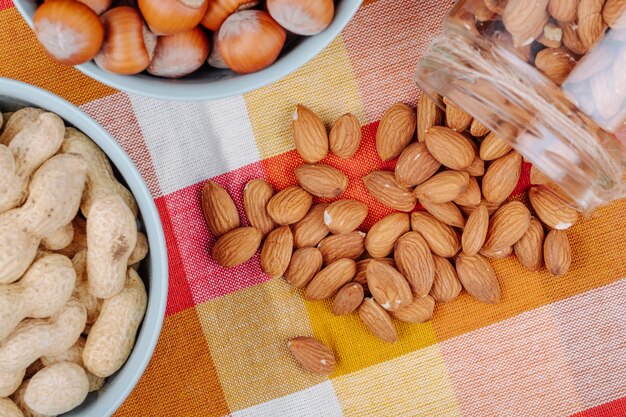 Vista superior de nueces, cacahuetes, avellanas en tazones y almendras esparcidas de un frasco de vidrio sobre una servilleta de tela escocesa