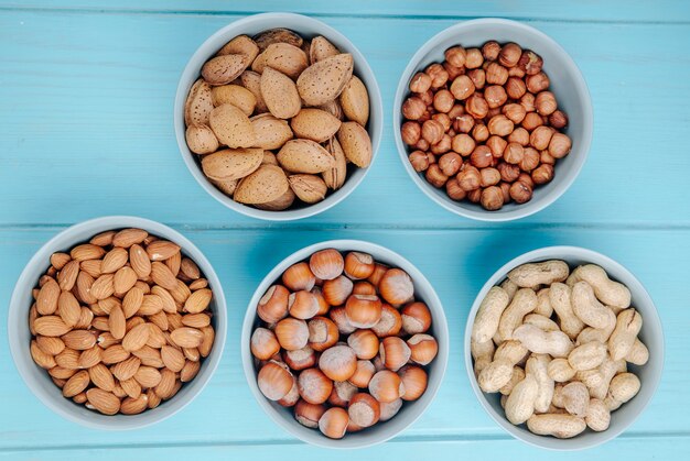 Vista superior de nueces mixtas con cáscara y sin cáscara en cuencos, almendras, avellanas y cacahuetes sobre fondo azul.