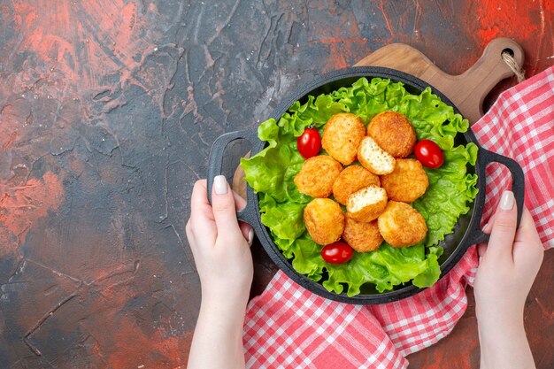 Vista superior de nuggets de pollo lechuga tomates cherry en sartén en manos femeninas en el espacio libre de la pared de color rojo oscuro