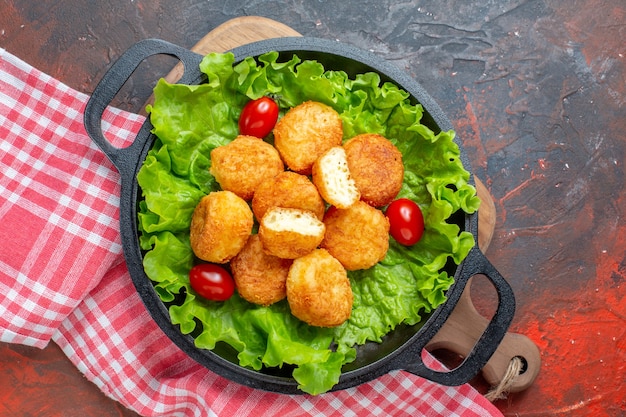 Vista superior de nuggets de pollo, lechuga, tomates cherry en una sartén en la tabla de cortar en la pared de color rojo oscuro.