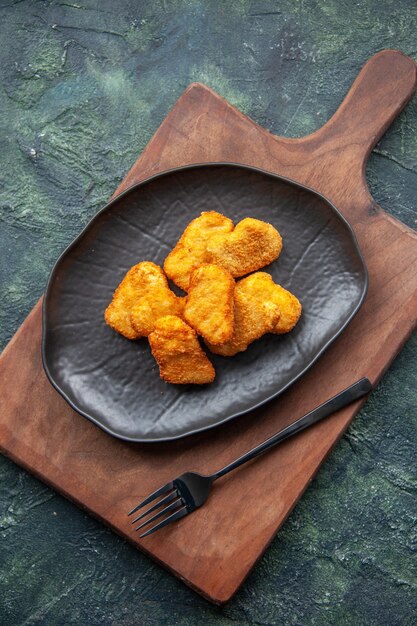 Vista superior de nuggets de pollo en un plato negro y tenedor sobre tabla de cortar de madera sobre superficie oscura