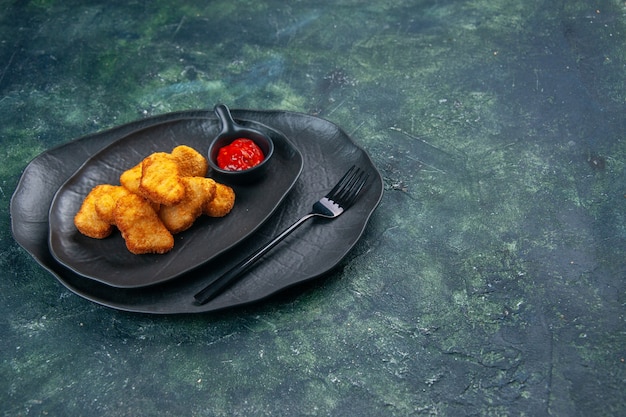 Vista superior de nuggets de pollo y tenedor de salsa de tomate en placas negras en el lado derecho sobre una superficie oscura