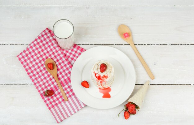 Vista superior de obleas de arroz blanco en un plato con mantel de cuadros rojos, fresas, cucharas de madera y leche en la superficie de la tabla de madera blanca. horizontal