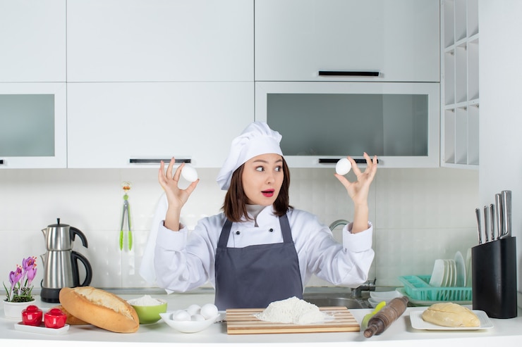 Foto gratuito vista superior de la ocupada chef femenina en uniforme de pie detrás de la mesa con alimentos de tabla de cortar con huevos en la cocina blanca