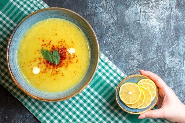 Vista superior de una olla azul con sabrosa sopa servida con menta y mano tomando limón picado sobre fondo azul.