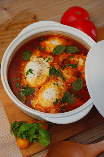 Vista superior de una olla blanca de shakshuka con huevos y hierbas frescas sobre una superficie de madera