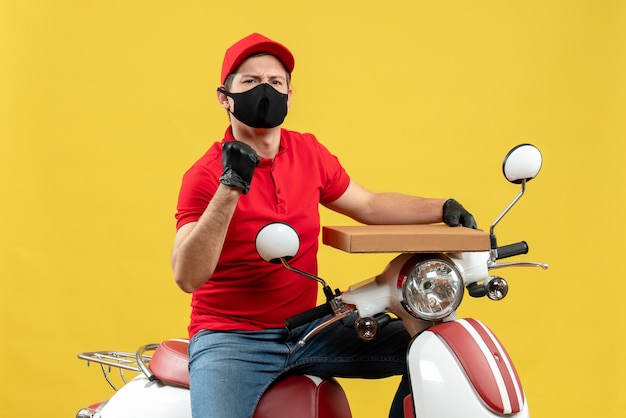 Vista superior del orgulloso mensajero emocional ambicioso vestido con blusa roja y guantes de sombrero en máscara médica sentado en scooter mostrando orden