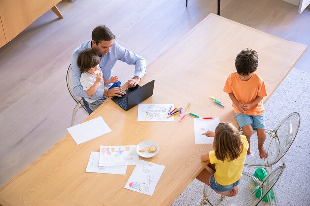 Vista superior del padre con niños sentados en la mesa. Hermano y hermana dibujando garabatos con marcadores. Papá de mediana edad trabajando en una computadora portátil y sosteniendo a su pequeño hijo. Concepto de infancia, fin de semana y tiempo en familia.