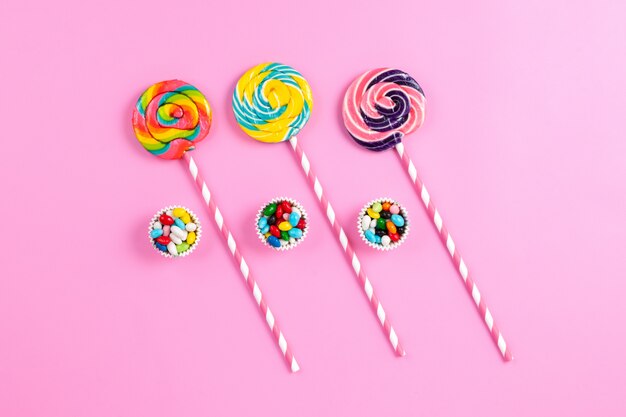 Una vista superior de paletas dulces de colores en los palitos de azúcar de color rosa-blanco y caramelos multicolores en el arco iris de cumpleaños de fondo rosa
