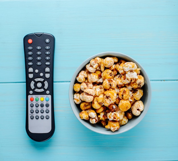 Vista superior de palomitas de caramelo dulce con control remoto de tv sobre fondo azul.