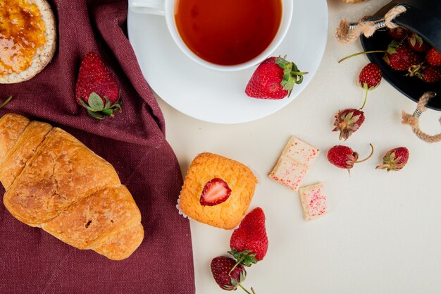 Vista superior de pan crujiente crujiente sobre tela y taza de té con fresas y magdalena con chocolate blanco sobre superficie blanca