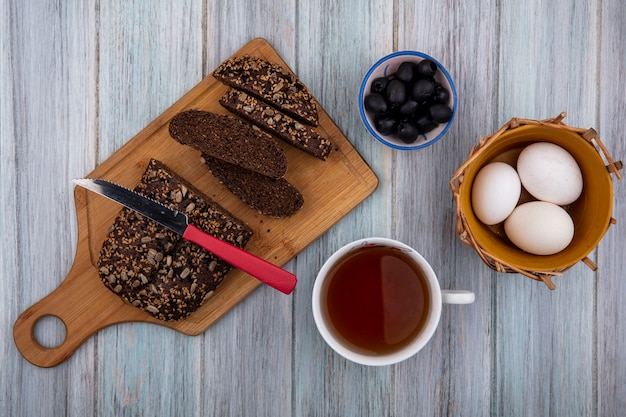 Vista superior de pan negro con cuchillo a bordo con taza de té huevos de gallina y aceitunas sobre fondo gris