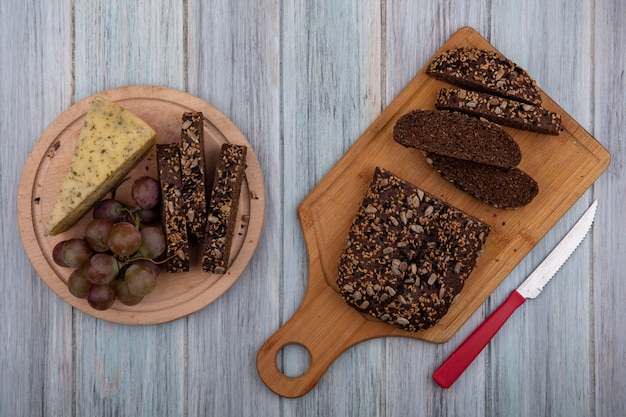 Vista superior de pan negro con un cuchillo en una tabla con uvas y queso en un soporte sobre un fondo gris