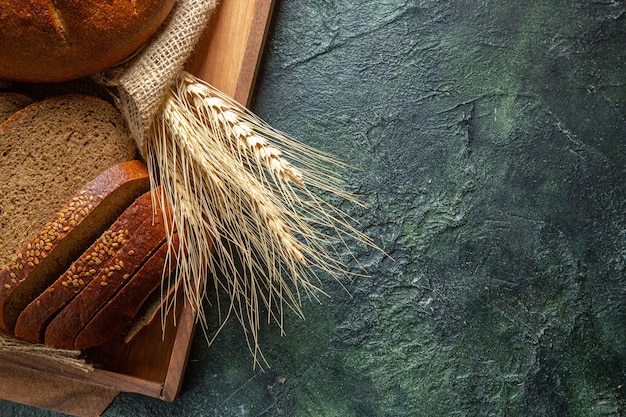 Foto gratuita vista superior del pan negro fresco entero y cortado sobre una toalla en una bandeja de madera marrón sobre fondo oscuro de colores mezcla