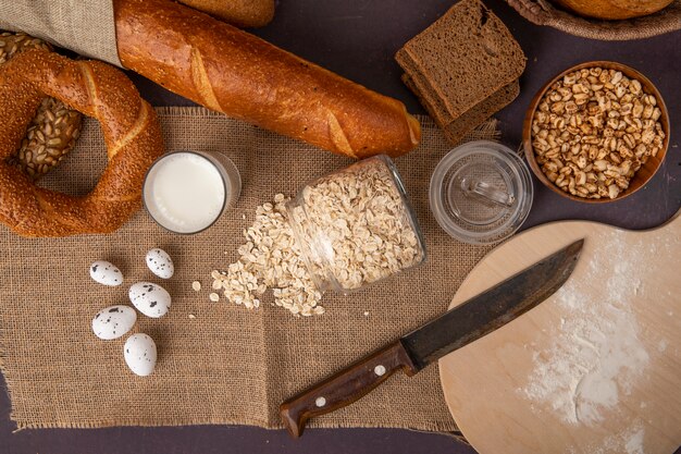 Vista superior de panes como bagel centeno y baguette con leche, copos de avena, huevos, callos y cuchillo sobre tela de saco sobre fondo marrón