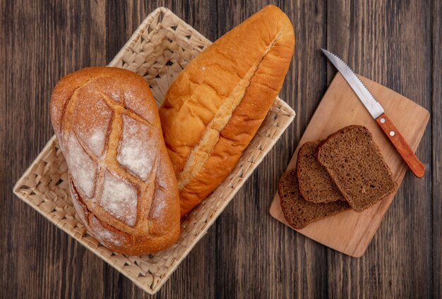 Vista superior de panes como baguette crujiente y vietnamita en la canasta y pan de centeno en rodajas con un cuchillo en la tabla de cortar sobre fondo de madera