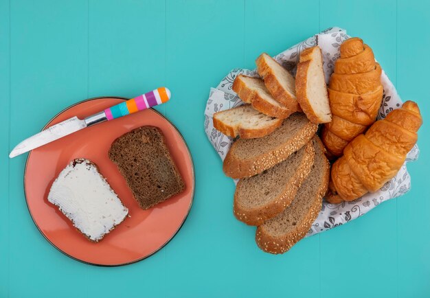 Vista superior de panes como baguette de mazorca sin semillas en rodajas y croissant en un tazón y pan de centeno untado con queso con un cuchillo en un plato sobre fondo azul.