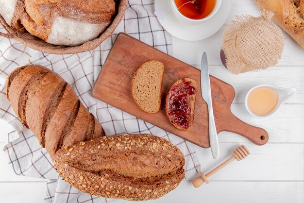 Vista superior de panes como baguette vietnamita negra con semillas negras pan negro de mazorca y centeno con mermelada y cuchillo en la tabla de cortar con mantequilla de té en la mesa de madera