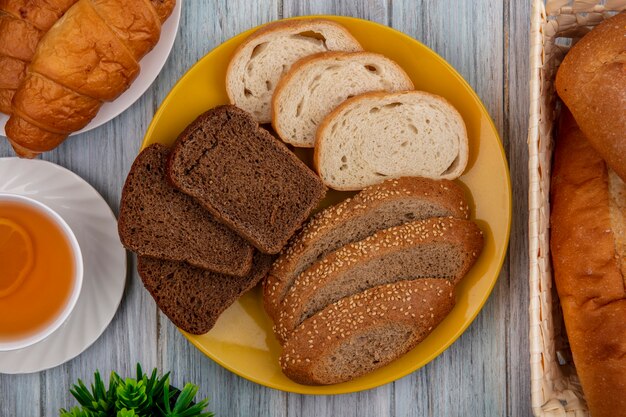 Vista superior de los panes como croissant de baguette de croissant de mazorca marrón sin semillas en rodajas de centeno y blancos en platos y en la cesta con toddy caliente sobre fondo de madera