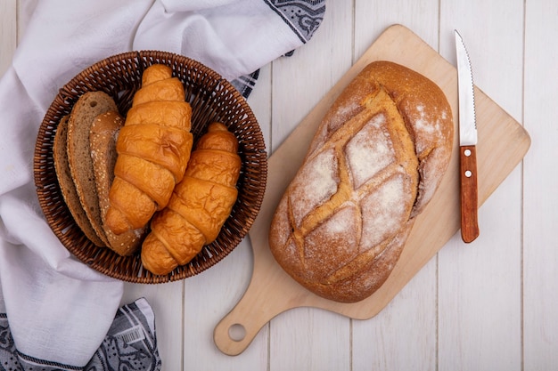 Vista superior de panes como croissant y rebanadas de pan de mazorca marrón sin semillas en la canasta sobre tela y pan crujiente sobre tabla de cortar sobre fondo de madera