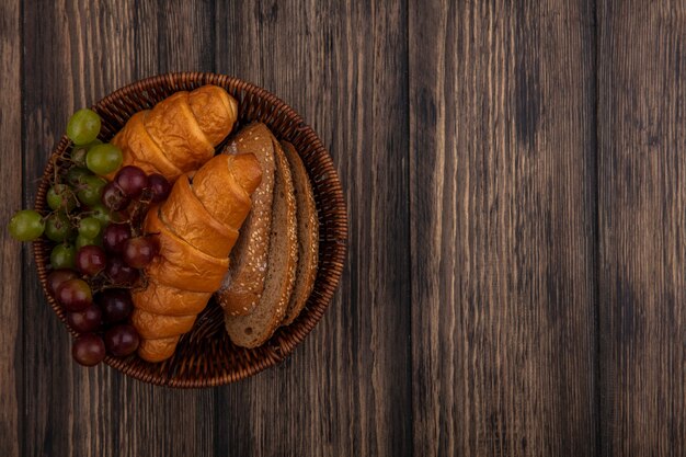 Vista superior de panes como croissants y rebanadas de pan de mazorca marrón sin semillas con uva en la canasta sobre fondo de madera con espacio de copia