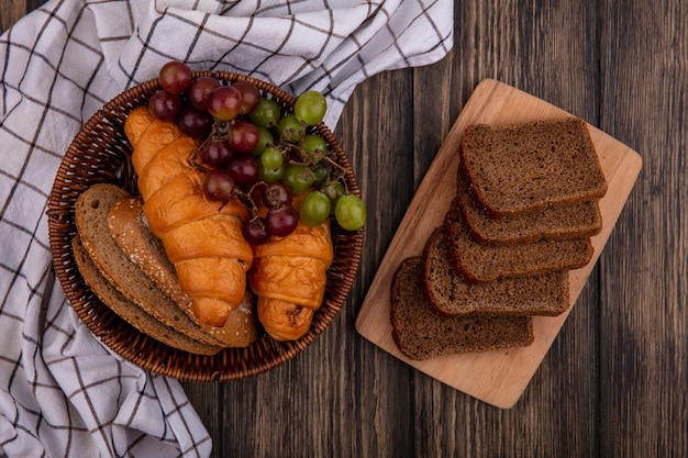 Vista superior de panes como croissants y rebanadas de pan de mazorca marrón sin semillas con uva en la canasta sobre tela a cuadros y rebanadas de pan de centeno en la tabla de cortar sobre fondo de madera