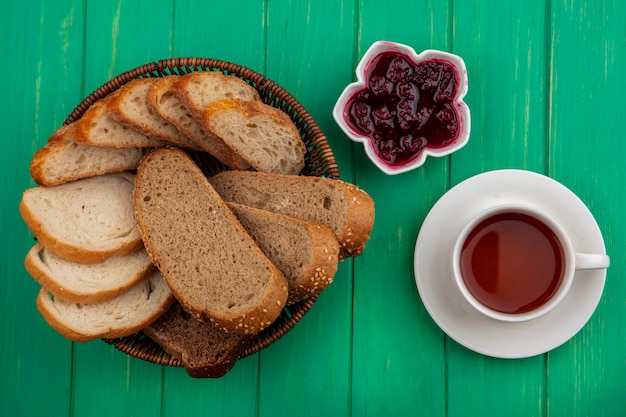 Vista superior de panes como rebanadas de mazorca marrón y baguette en canasta y mermelada de frambuesa en un tazón con taza de té sobre fondo verde