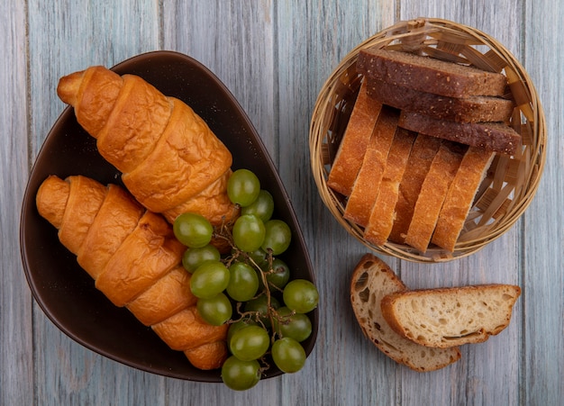 Vista superior de panes croissants en rodajas de centeno y crujientes en un tazón y en la canasta con uva sobre fondo de madera