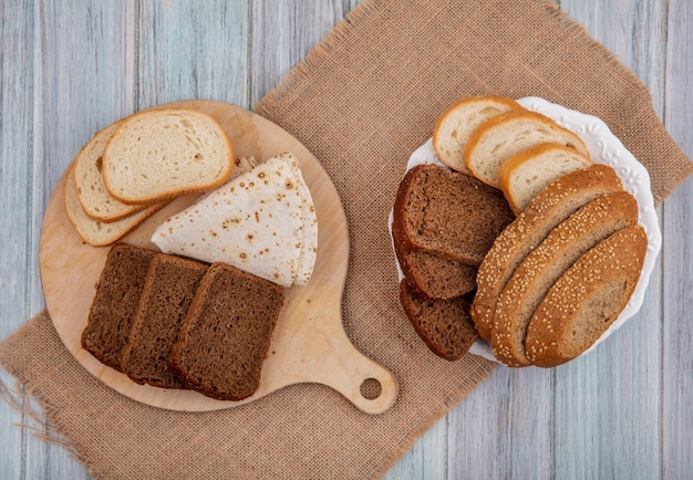 Vista superior de los panes en rodajas de centeno blanco sembrado mazorca marrón y pan plano en la tabla de cortar y en el plato sobre tela de saco sobre fondo de madera