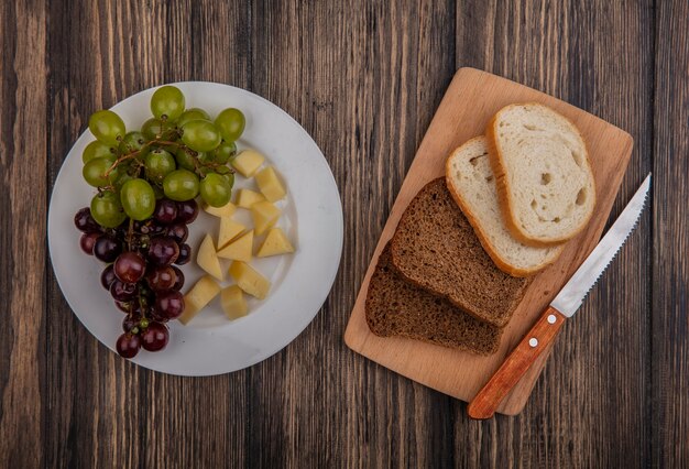 Vista superior de panes en rodajas de centeno y blancos con un cuchillo en la tabla de cortar y un plato de uva y queso sobre fondo de madera