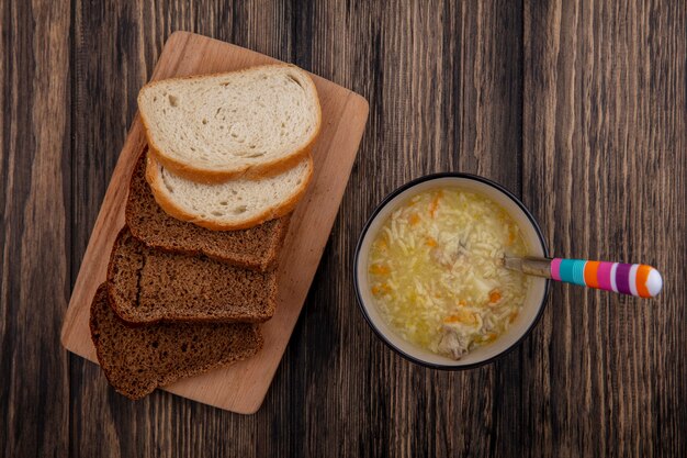 Vista superior de panes en rodajas de centeno y blancos sobre tabla de cortar y tazón de sopa de pollo orzo con cuchara sobre fondo de madera