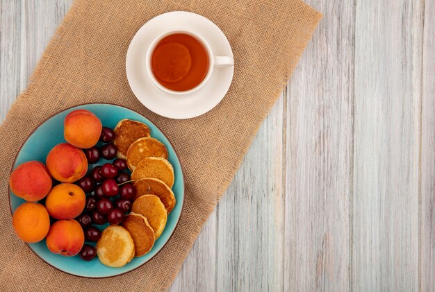 Vista superior de panqueques con cerezas y albaricoques en plato y taza de té en platillo sobre tela de saco y fondo de madera con espacio de copia