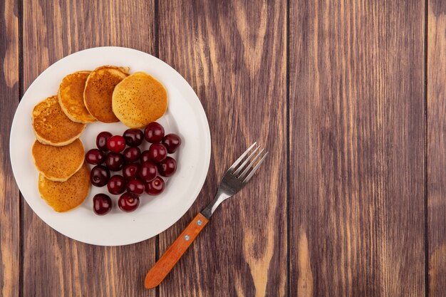 Vista superior de panqueques con cerezas en plato y tenedor sobre fondo de madera con espacio de copia