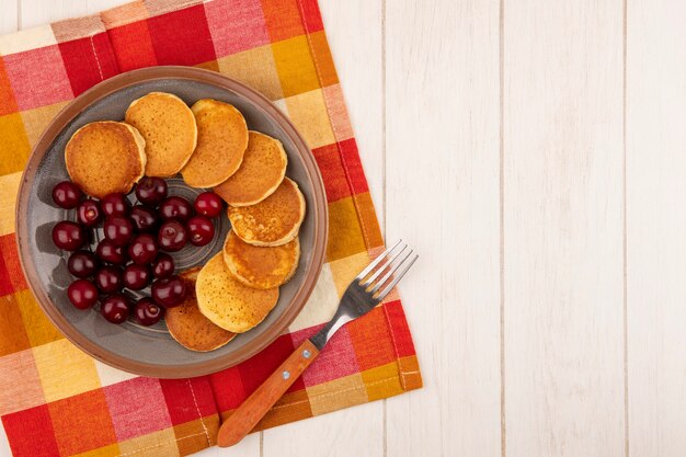 Vista superior de panqueques con cerezas en plato y tenedor sobre tela a cuadros y fondo de madera con espacio de copia