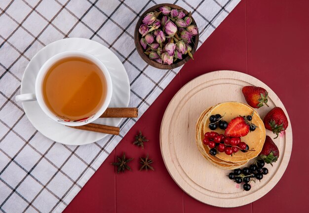 Vista superior de panqueques con grosellas rojas y negras y fresas en una bandeja con una taza de té y canela sobre un fondo rojo.