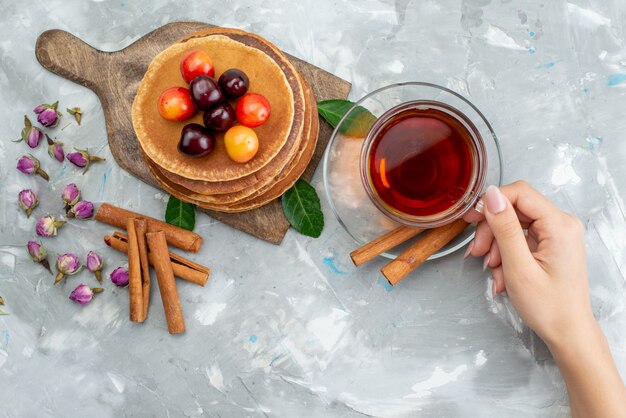 Una vista superior panqueques redondos horneados y deliciosos con cerezas y té femenino en la mesa de luz pastel de frutas
