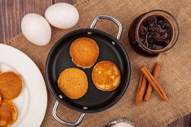 Vista superior de panqueques en sartén y en plato y tarro de mermelada de fresa con canela y huevos en tela de saco sobre fondo de madera