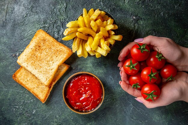 Vista superior de papas fritas con tostadas y tomates cherry rojos en manos femeninas sobre superficie oscura