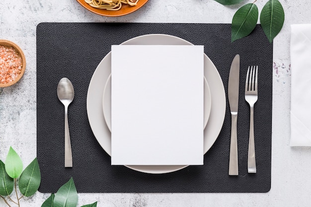 Vista superior del papel de menú en blanco en un plato con cubiertos y hojas