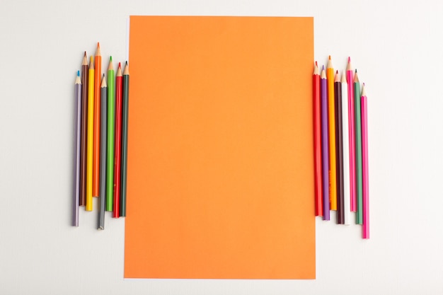 Vista superior de papel naranja en blanco con lápices de colores sobre superficie blanca